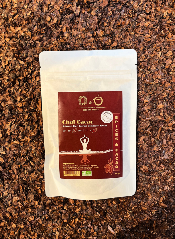 Un sachet d'infusion au cacao cacao en papier blanc avec une étiquette rouge indiquant Chaï Cacao avec l'inscription "Origines Chocolate Maker Bordeaux" posé dans du grué de cacao. 