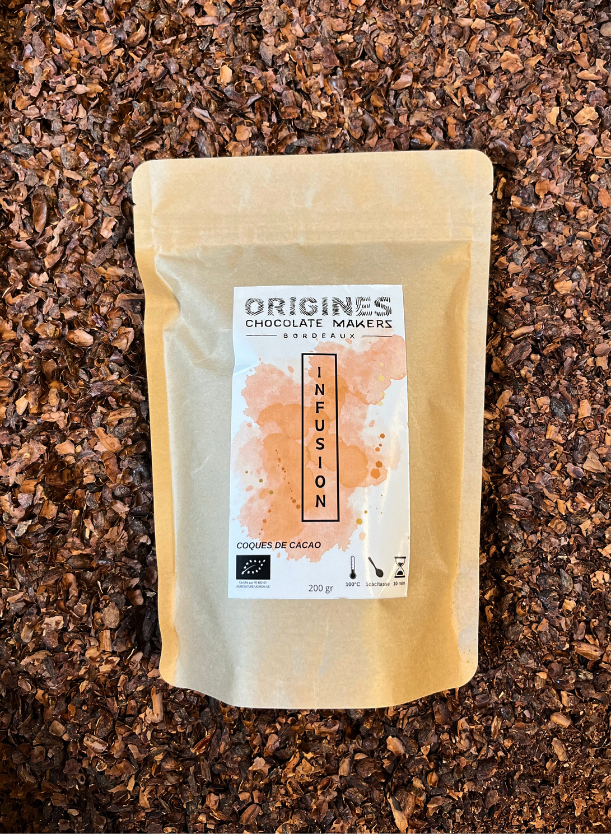 Un sachet d'infusion au cacao cacao en papier kraft avec l'inscription "Origines Chocolate Maker Bordeaux" posé dans du grué de cacao. 