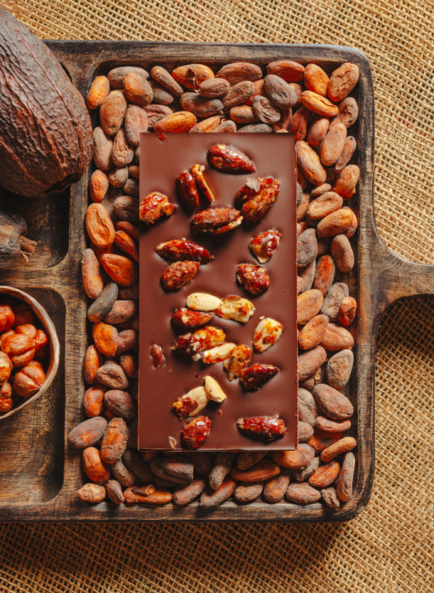 Tablette de chocolat noir au amandes de Valence Origines Chocolate Makers 70 % de cacao provenant du Pérou, entourée de fèves de cacao, de cabosses et d'un bol contenant des amandes sur un fond en toile de jute.