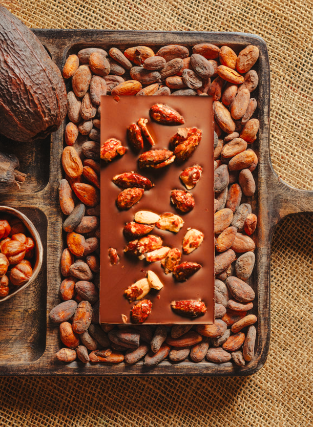 Tablette de chocolat au lait au amandes de Valence Origines Chocolate Makers,38 % de cacao de São Tomé entourée de fèves de cacao, de cabosses et d'un bol contenant des amandes sur un fond en toile de jute. 