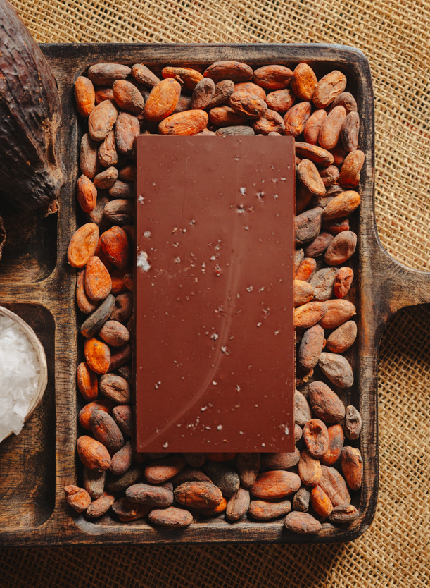 Tablette de chocolat noir à la fleur de sel du bassin d'Arcachon Origines Chocolate Makers, 70 % de cacao provenant du Pérou, entourée de fèves de cacao, de cabosses et d'un bol contenant du sel sur un fond en toile de jute.