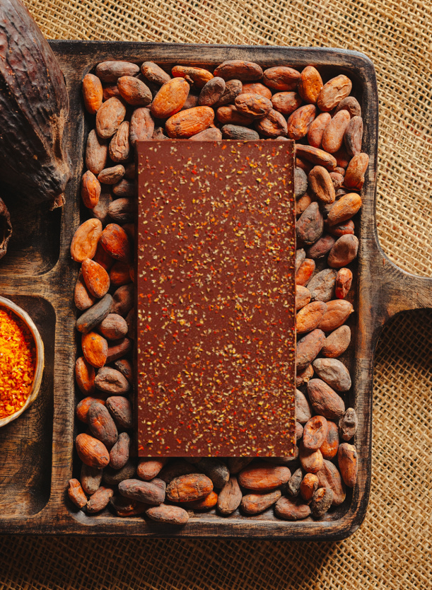 Tablette de chocolat noir au piment d'Espelette Origines Chocolate Makers 70 % de cacao provenant du Pérou, entourée de fèves de cacao, de cabosses et d'un bol contenant du piment d'Espelette sur un fond en toile de jute.