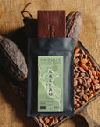 Tablette de chocolat noir Origines Chocolate Makers, emballée dans un sachet noir avec une étiquette verte indiquant 85% de cacao provenant du Pérou, entourée de fèves de cacao et de cabosses sur un fond en toile de jute.