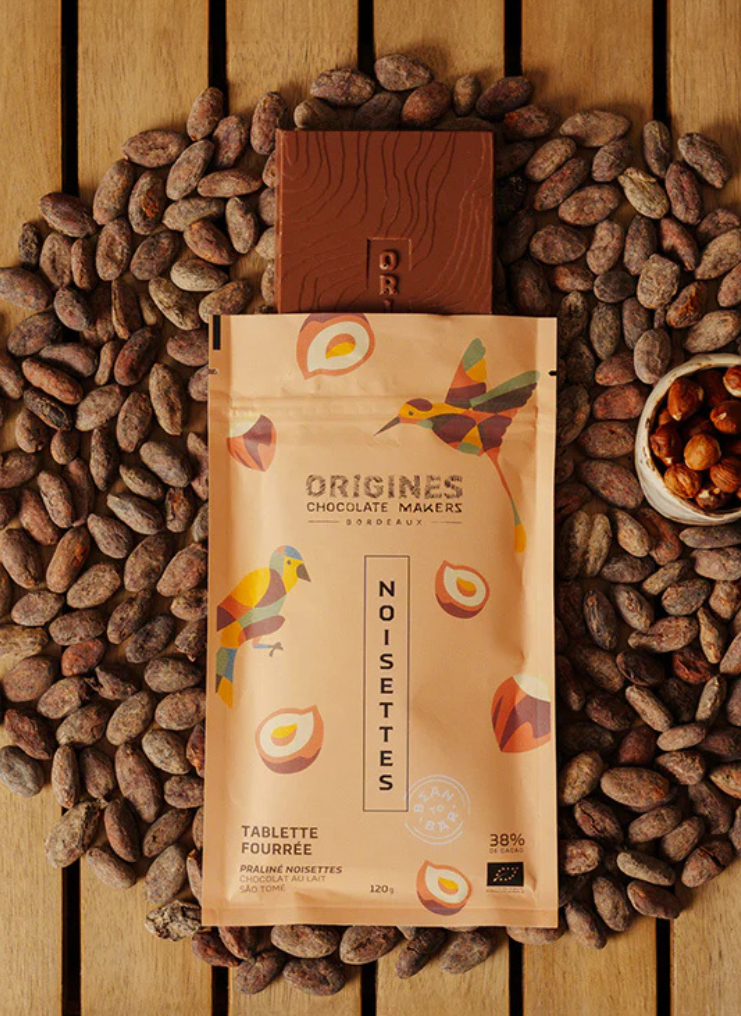Tablette de chocolat noir Origines Chocolate Makers à la noisette, emballée dans un sachet orange orné de toucans colorés, entourée de fèves de cacao et d'un petit bol de noisettes sur une surface en bois.