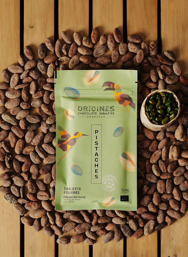 Tablette de chocolat noir Origines Chocolate Makers à la pistache, emballée dans un sachet vert orné de toucans colorés, entourée de fèves de cacao et d'un petit bol de pistaches sur une surface en bois.