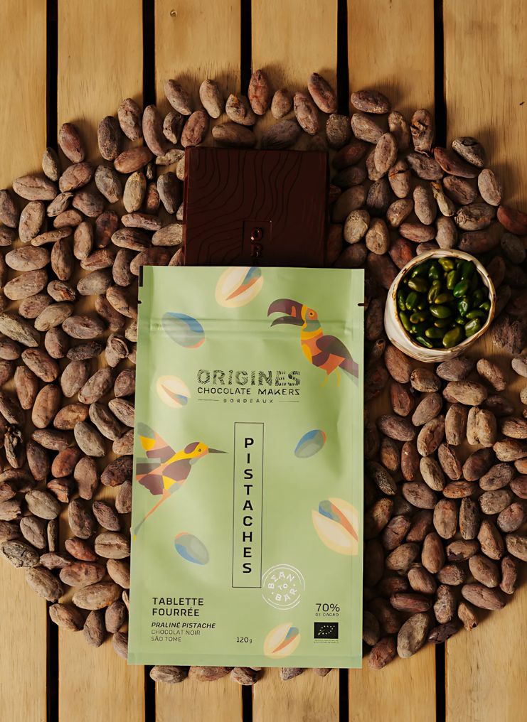 Tablette de chocolat noir Origines Chocolate Makers à la pistache, emballée dans un sachet vert orné de toucans colorés, entourée de fèves de cacao et d'un petit bol de pistaches sur une surface en bois.