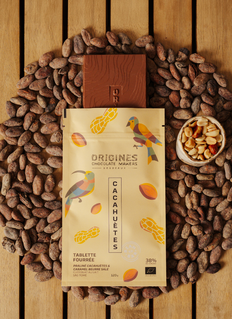 Tablette de chocolat noir Origines Chocolate Makers à la cacahuète, emballée dans un sachet jaune orné de toucans colorés, entourée de fèves de cacao et d'un petit bol de cacahuètes sur une surface en bois.