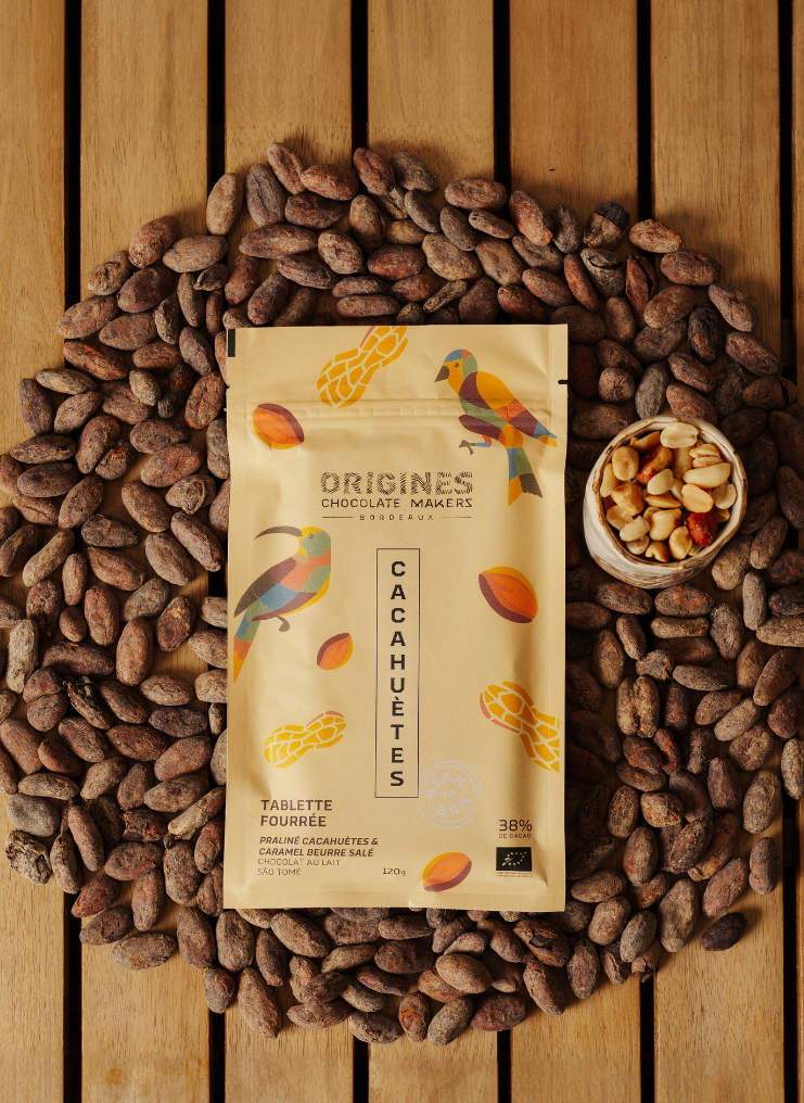 Tablette de chocolat noir Origines Chocolate Makers à la cacahuète, emballée dans un sachet jaune orné de toucans colorés, entourée de fèves de cacao et d'un petit bol de cacahuètes sur une surface en bois.