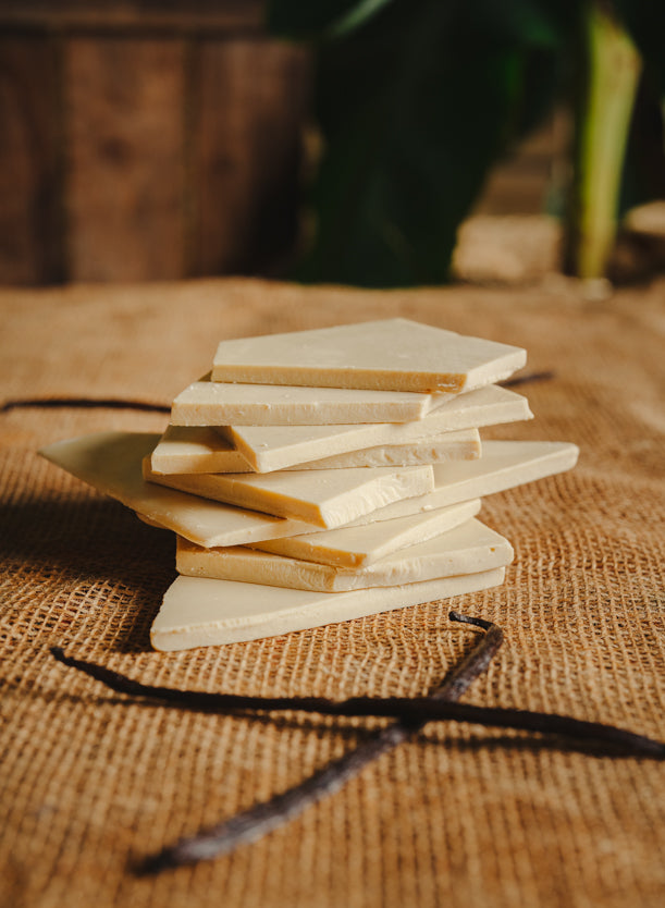 Morceaux de chocolat blanc à la vanille empilés sur une toile de jute, avec des gousses de vanille dispersées autour, en arrière-plan naturel.
