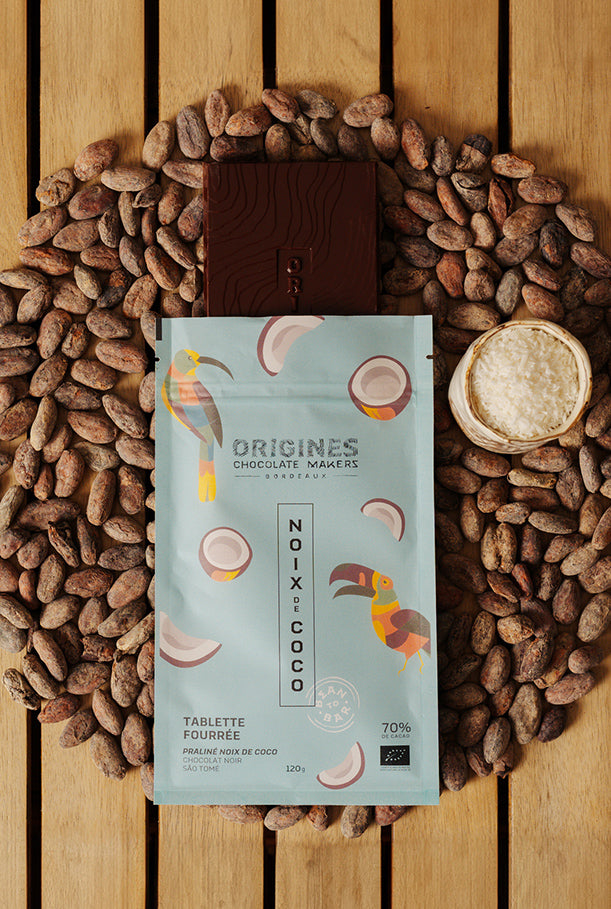 Tablette de chocolat noir Origines Chocolate Makers à la noix de coco, emballée dans un sachet bleuorné de toucans colorés, entourée de fèves de cacao et d'un petit bol de noix de coco sur une surface en bois.