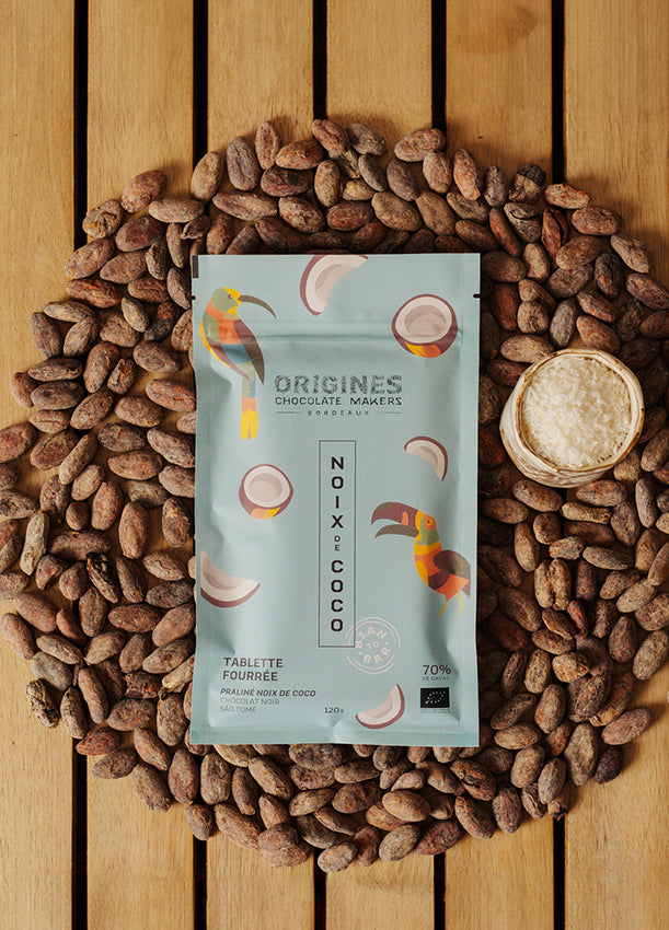 Tablette de chocolat noir Origines Chocolate Makers à la noix de coco, emballée dans un sachet bleuorné de toucans colorés, entourée de fèves de cacao et d'un petit bol de noix de coco sur une surface en bois.