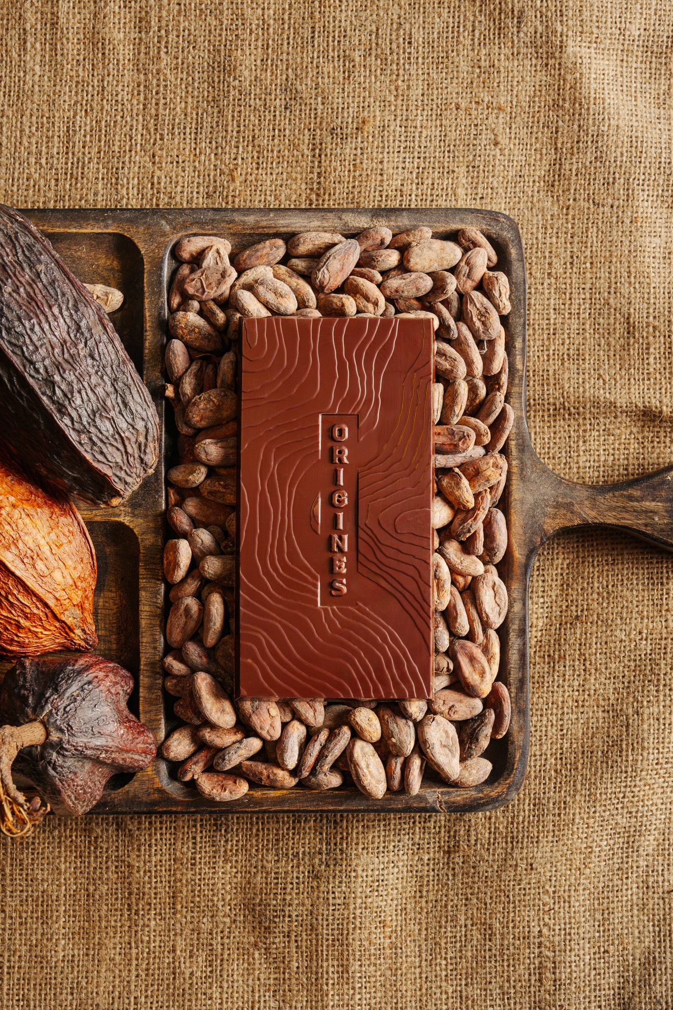 Tablette de chocolat noir Origines Chocolate Makers 70 % de cacao provenant de la coopérative Cabruca à Bahia, Brésil, entourée de fèves de cacao et de cabosses sur un fond en toile de jute.